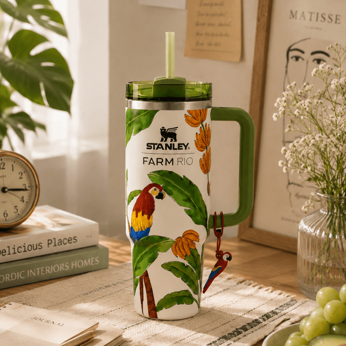 A Stanley Farm Rio insulated tumbler with a green handle and straw, decorated with colorful parrots and tropical leaves, set on a wooden table in a styled home environment.