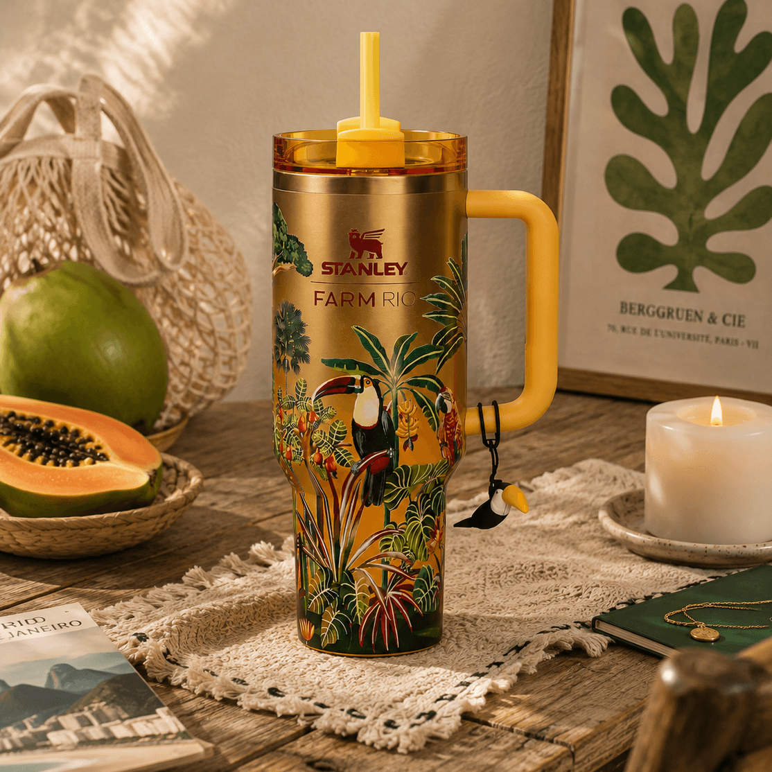 A Stanley insulated tumbler with a yellow handle and lid, decorated with tropical toucan and palm tree artwork, set on a rustic wooden table with a candle, fruit, and framed wall art in the background.