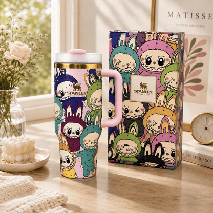 A Stanley London travel mug with a pink handle and lid, featuring a colorful cartoon rabbit character print, shown next to its matching illustrated box on a wooden table.