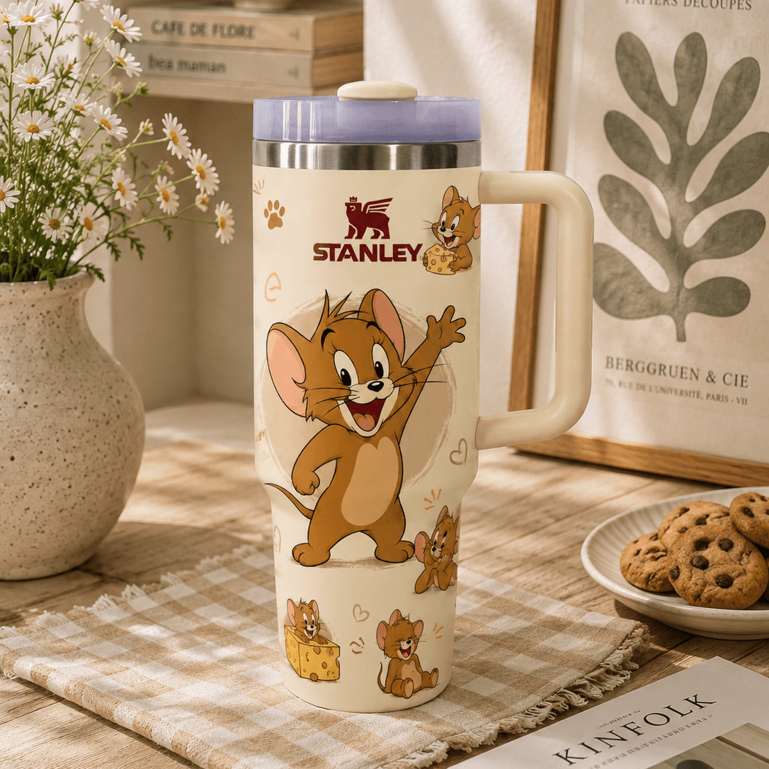 A Stanley branded insulated travel mug with a cartoon Jerry design, styled on a wooden tabletop with a vase of daisies, cookies, a book, and framed wall art in a warm indoor setting.