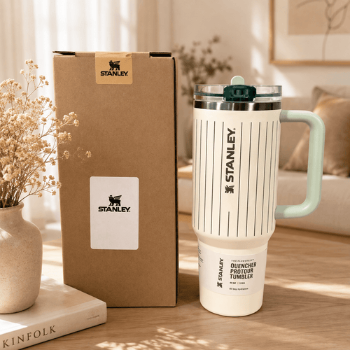 A Stanley duquencher pourtour tumbler in off-white with black vertical stripes and a mint green handle, shown next to its brown cardboard packaging box on a wooden table.