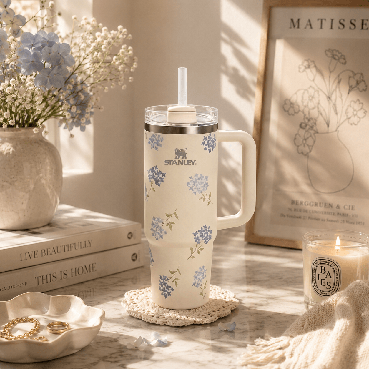 A cream-colored insulated tumbler with a floral pattern in blue and green, featuring the STANLEY logo, sits on a marble surface in a beautifully styled room setting with a candle, books, and dried flowers.