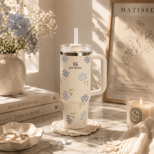 A cream-colored insulated tumbler with a floral pattern in blue and green, featuring the STANLEY logo, sits on a marble surface in a beautifully styled room setting with a candle, books, and dried flowers.