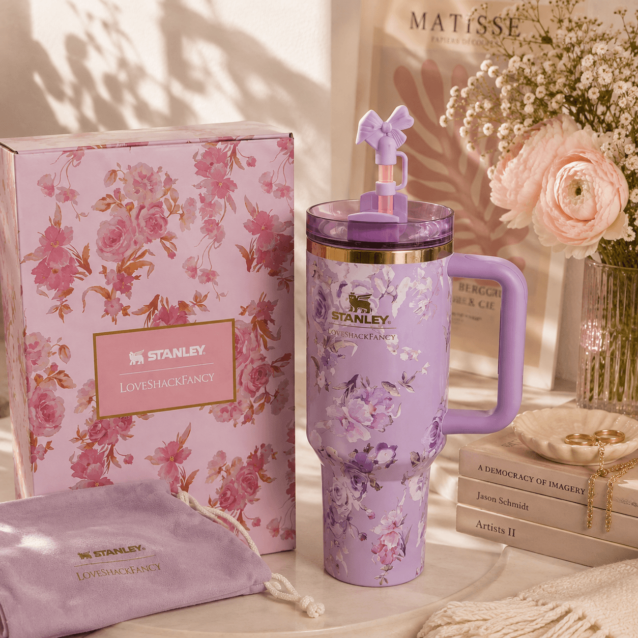 A lavender Stanley travel mug with a floral print, purple lid, and integrated handle, displayed next to its pink floral gift box and matching drawstring pouch on a styled tabletop with flowers and books.