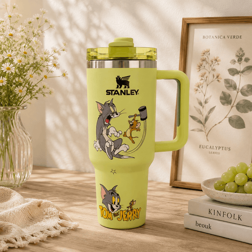 A light yellow-green Stanley tumbler with a handle, featuring Tom and Jerry cartoon graphics, set on a wooden table with a vase of white flowers, a framed botanical print, and a bowl of green grapes in the background.