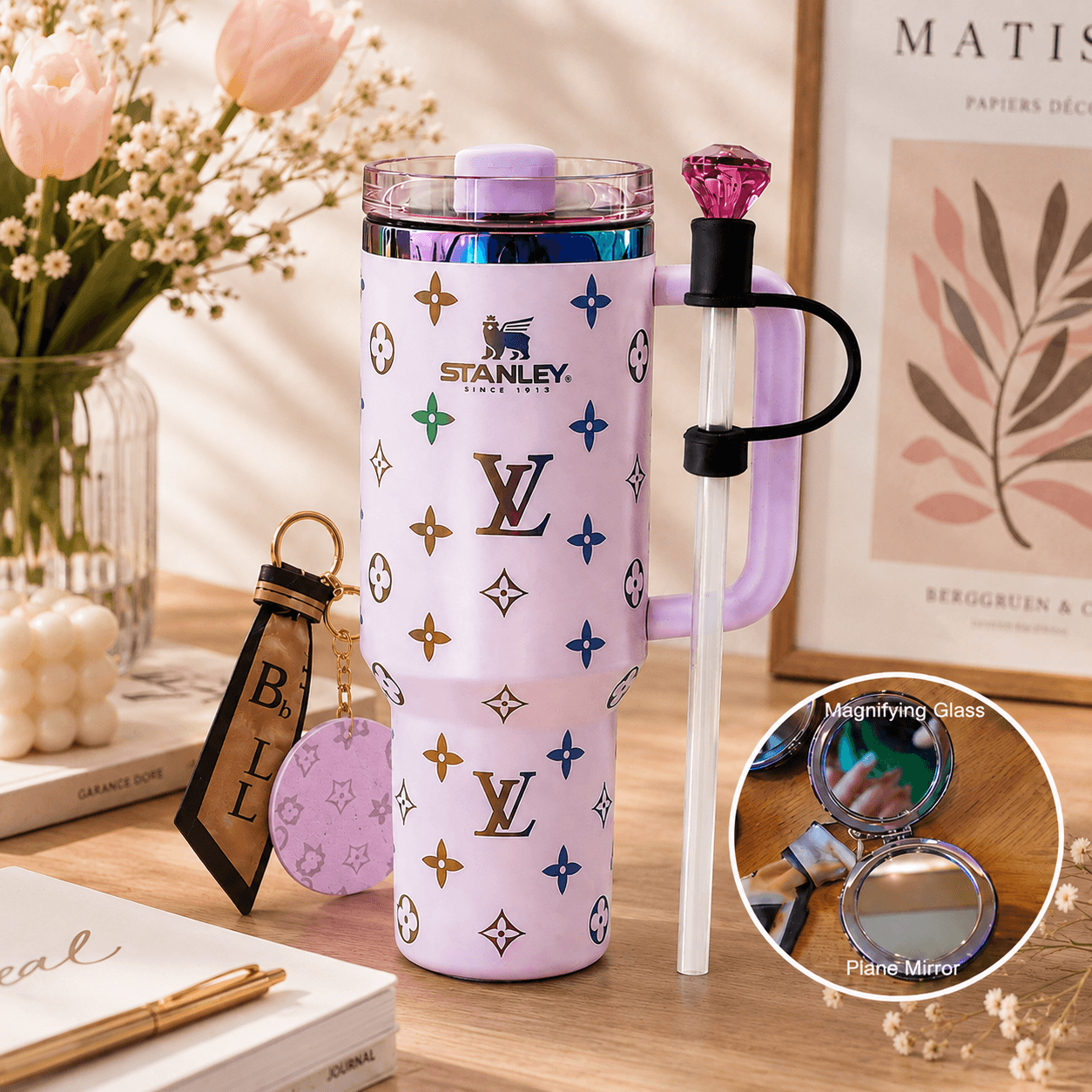 A pastel purple Stanley tumbler with a monogram pattern, a purple lid, and a clear handle with a purple ring and rhinestone accent, shown on a wooden table with decorative props and a circular inset showing mirror details.