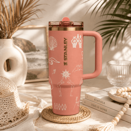 A pink Stanley travel tumbler with a handle and lid, decorated with white line-art illustrations, set on a woven coaster in a styled home environment with books, a hat, and palm leaves.