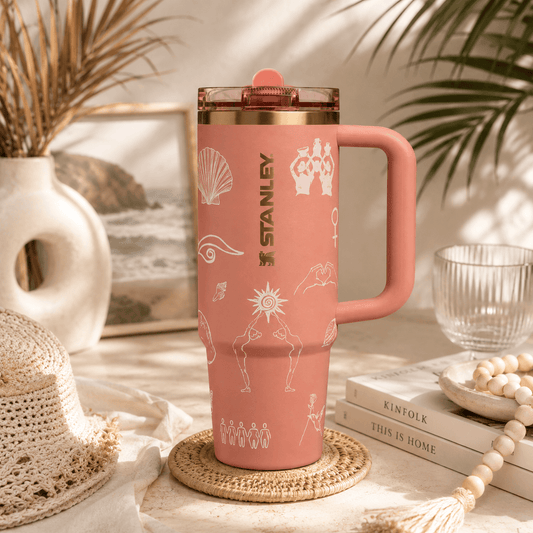 A pink Stanley travel tumbler with a handle and lid, decorated with white line-art illustrations, set on a woven coaster in a styled home environment with books, a hat, and palm leaves.