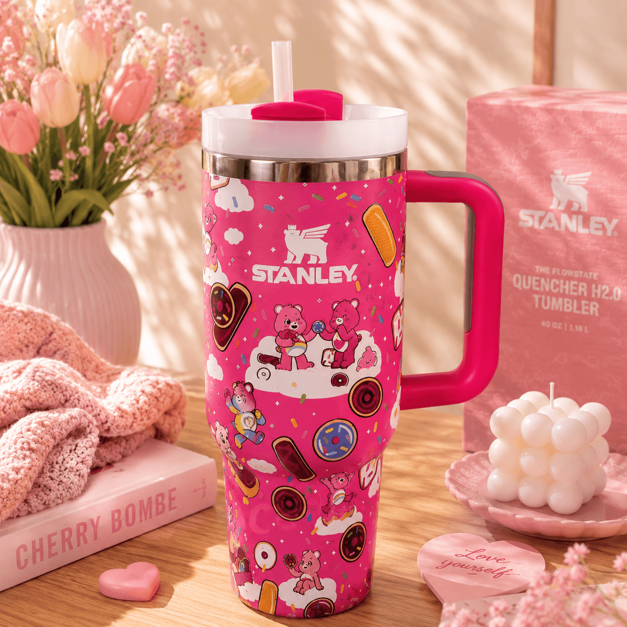 A pink Stanley tumbler with a handle and straw, decorated with cartoon teddy bears and candy motifs, displayed on a wooden table with a pink Stanley box, flowers, and small decorative items in a warm styled setting.