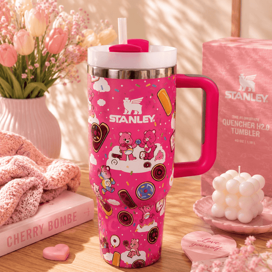 A pink Stanley tumbler with a handle and straw, decorated with cartoon teddy bears and candy motifs, displayed on a wooden table with a pink Stanley box, flowers, and small decorative items in a warm styled setting.