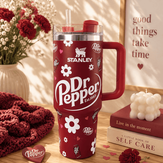 A red Stanley Dr Pepper insulated tumbler with a handle, clear lid, and straw, styled on a wooden surface with flowers, a knit blanket, books, and a framed quote in a warm indoor setting.