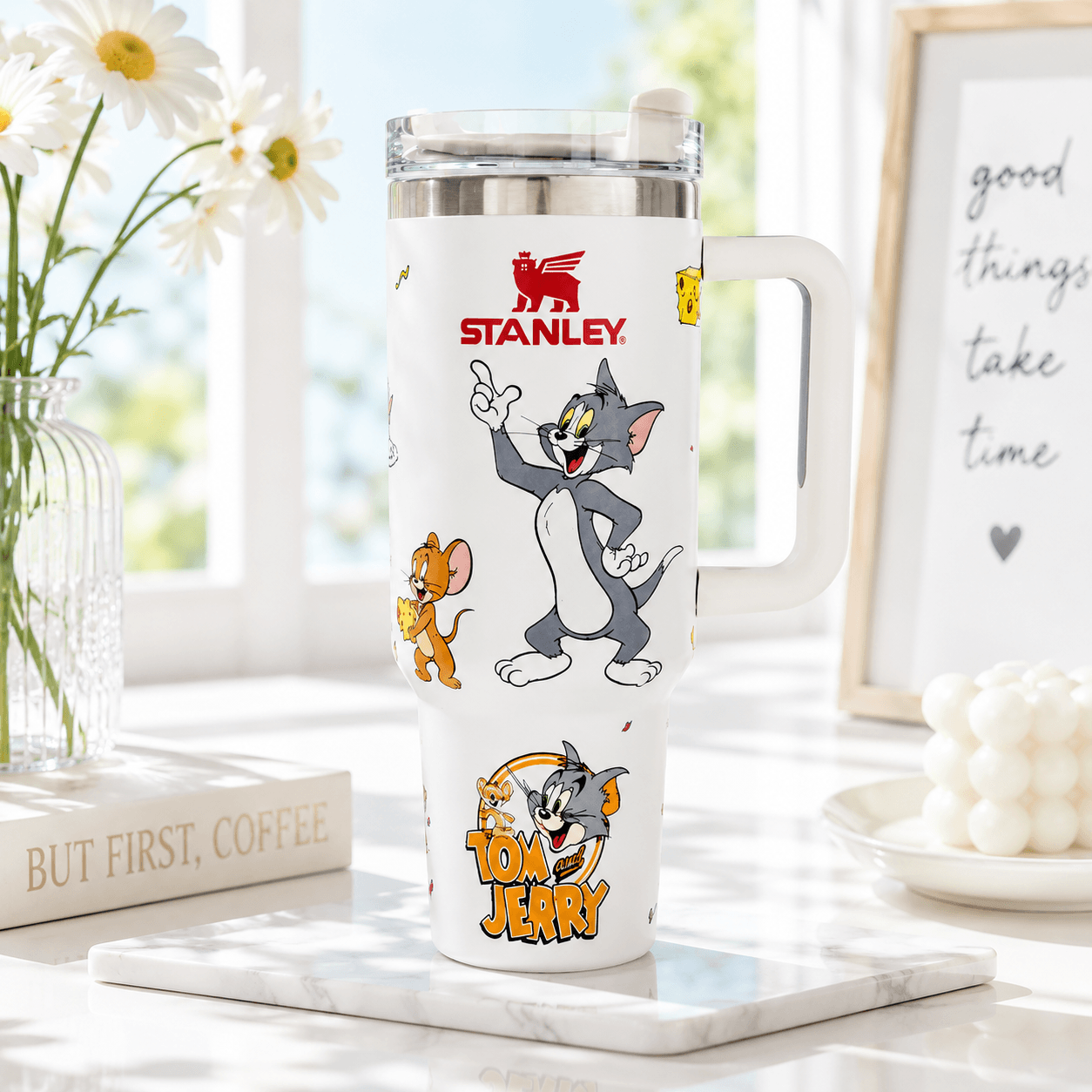 A white Stanley Tom and Jerry travel tumbler with a handle, shown on a marble tray in a bright, sunlit room. The tumbler features cartoon illustrations of Tom and Jerry and the red Stanley logo.