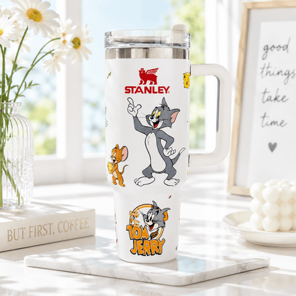 A white Stanley Tom and Jerry travel tumbler with a handle, shown on a marble tray in a bright, sunlit room. The tumbler features cartoon illustrations of Tom and Jerry and the red Stanley logo.