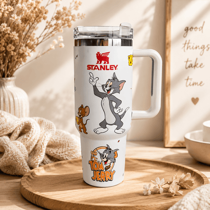 A white Stanley insulated tumbler with a handle and clear lid, decorated with Tom and Jerry cartoon graphics and logos, displayed on a wooden tray in a warm, styled interior setting.