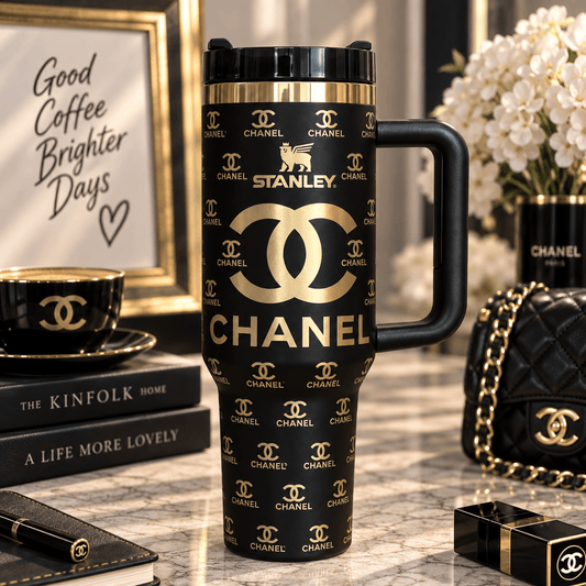 A black travel tumbler with gold Chanel and Stanley branding, styled on a marble surface with luxury accessories and framed decor in the background.