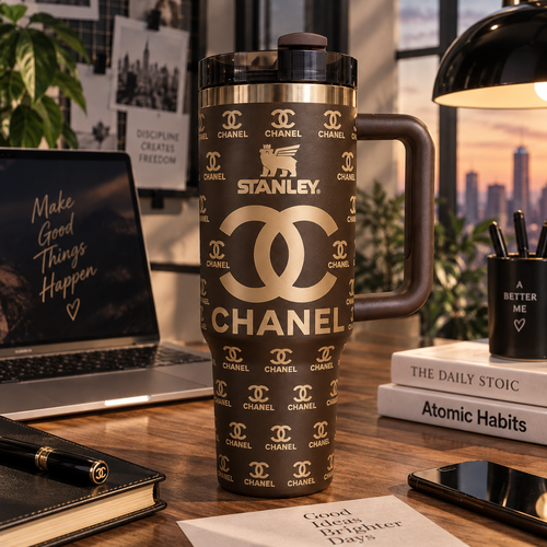 A dark brown Stanley travel mug with a gold Chanel logo pattern, set on a wooden desk surrounded by lifestyle props like a laptop, books, and a lamp.