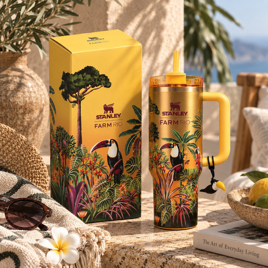 A Stanley Farm Rio insulated tumbler with a yellow handle and lid, shown beside its matching yellow box. The tumbler and packaging feature tropical artwork with toucans, palm leaves, and flowers.