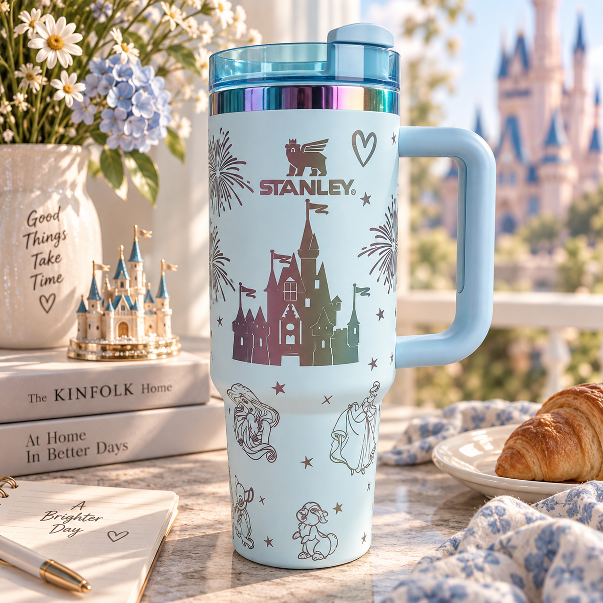 A light blue insulated travel tumbler with a handle and matching lid sits on a tabletop in a styled indoor setting. The tumbler is printed with castle and character graphics, including the name STANLEY above the castle.