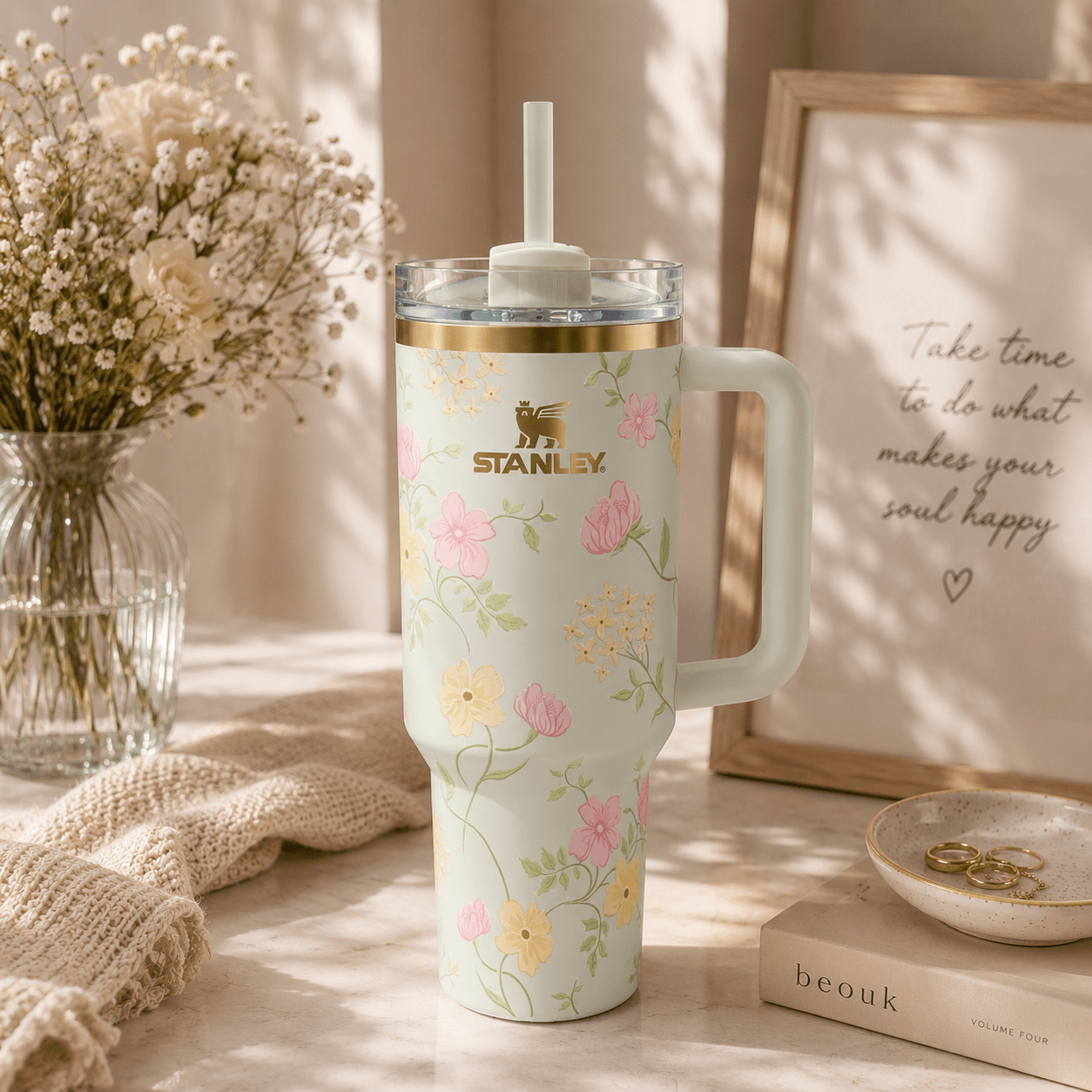 A Stanley handled tumbler with a clear lid and straw, decorated with a pastel floral print on a warm off-white background. The tumbler is styled on a light surface with a knit blanket, a vase of dried flowers, a framed quote, and a small dish in a softly lit indoor setting.