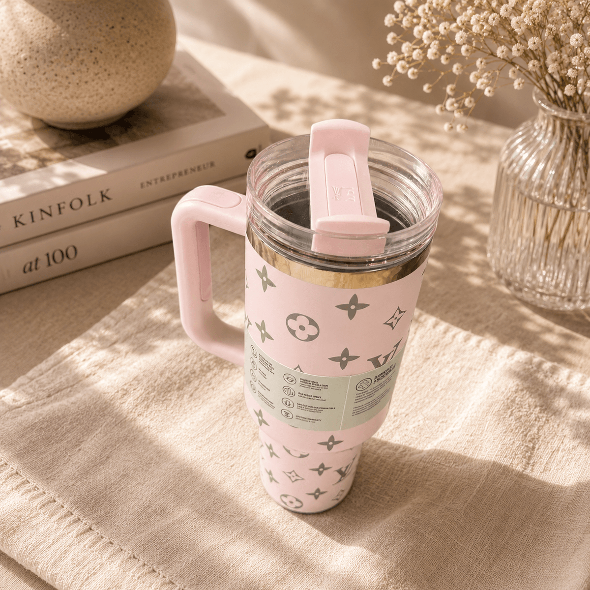 Pink travel mug with a pattern on a textured surface with books and a vase in the background