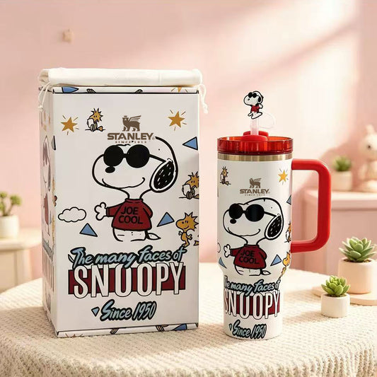 Stanley Snoopy insulated tumbler with a red handle and lid, shown beside its matching branded storage box on a soft pink tabletop. The tumbler and box feature Snoopy in sunglasses with 'JOE COOL' text and 'The many faces of SNOOPY Since 1959' artwork.