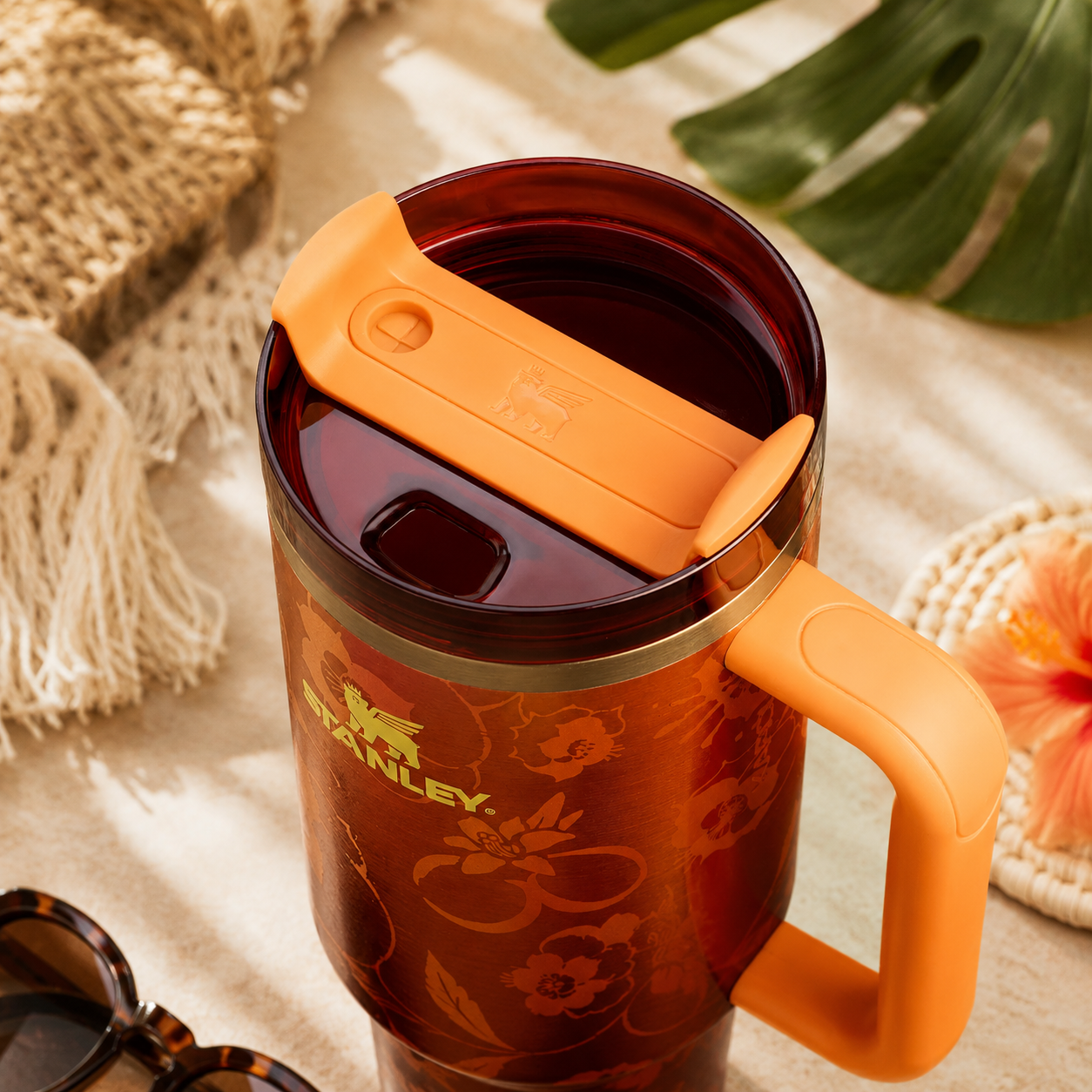 Stanley travel mug with orange handle and lid on a textured surface with leaves and sunglasses.