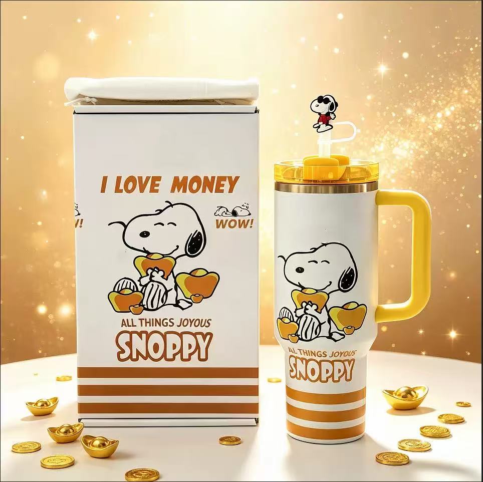 A white Snoopy tumbler with a yellow handle and lid, shown next to its matching white box packaging. The packaging and tumbler feature a cartoon print of Snoopy holding gold ingots with the text 'I LOVE MONEY', 'ALL THINGS JOYOUS', and 'SNOPPY'.